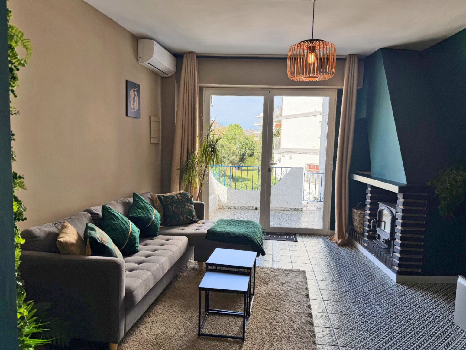 Woonkamer met bakstenen open haard, grijze bank met groene kussens en een terras met uitzicht.