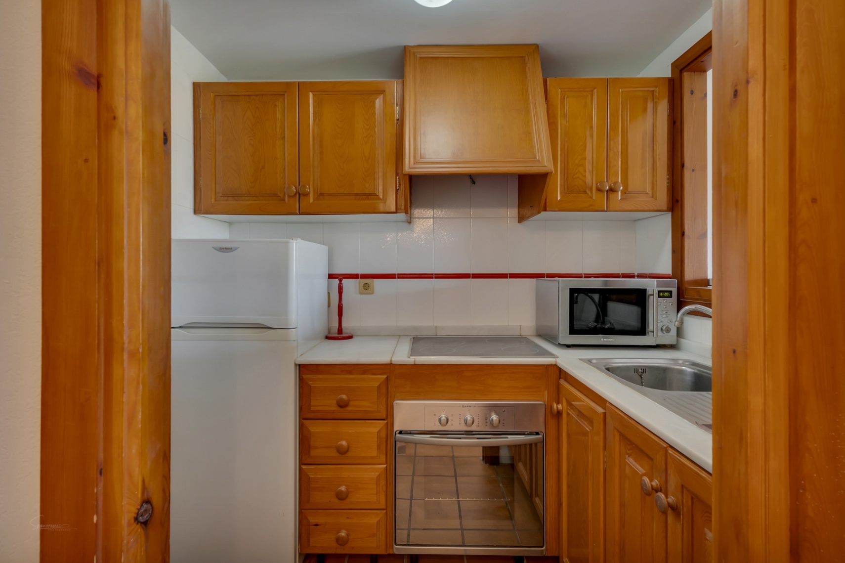 Compacte keuken met houten kasten, witte apparaten en een licht aanrecht.