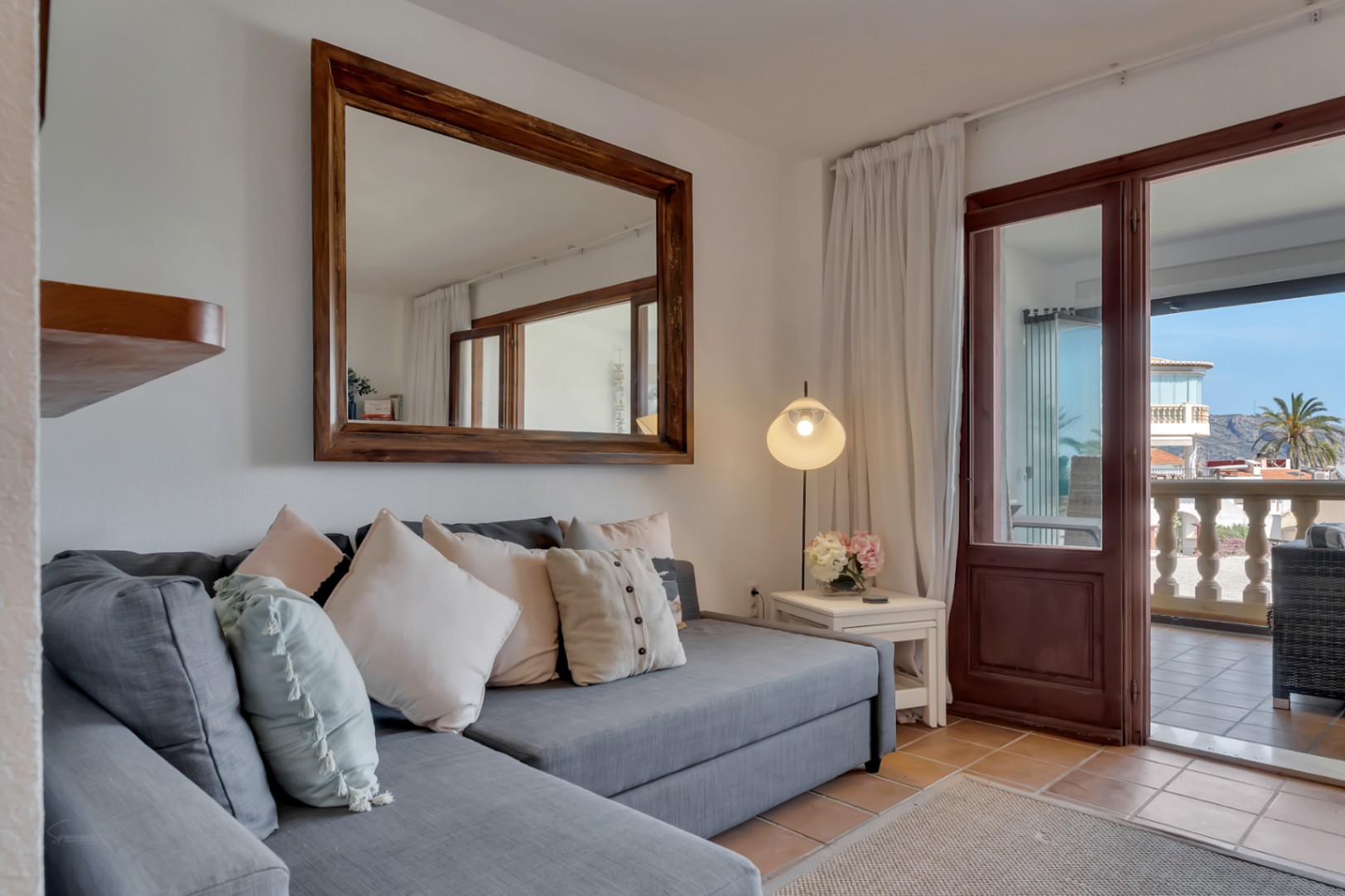 Woonkamer met grijze bank, houten spiegel en toegang tot terras met zeezicht in Jávea.