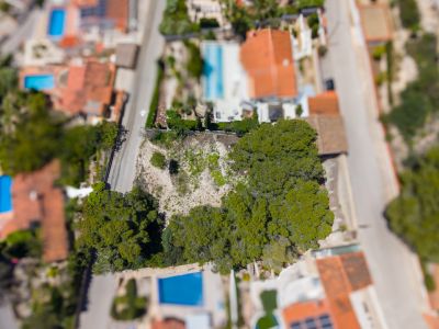 Luchtfoto van een onontwikkeld perceel met bomen, omringd door wegen en residentiële eigendommen met zwembaden en rode daken.