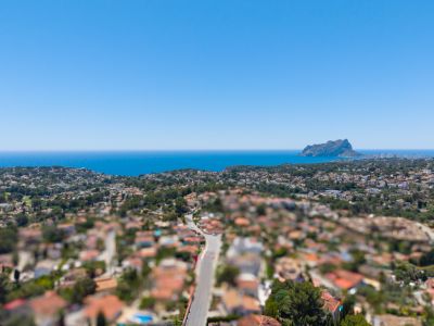 Panoramisch luchtfoto van de kust van Benissa, met de Middellandse Zee, de Peñón de Ifach en een residentiële wijk met weelderige begroeiing onder een helderblauwe lucht.