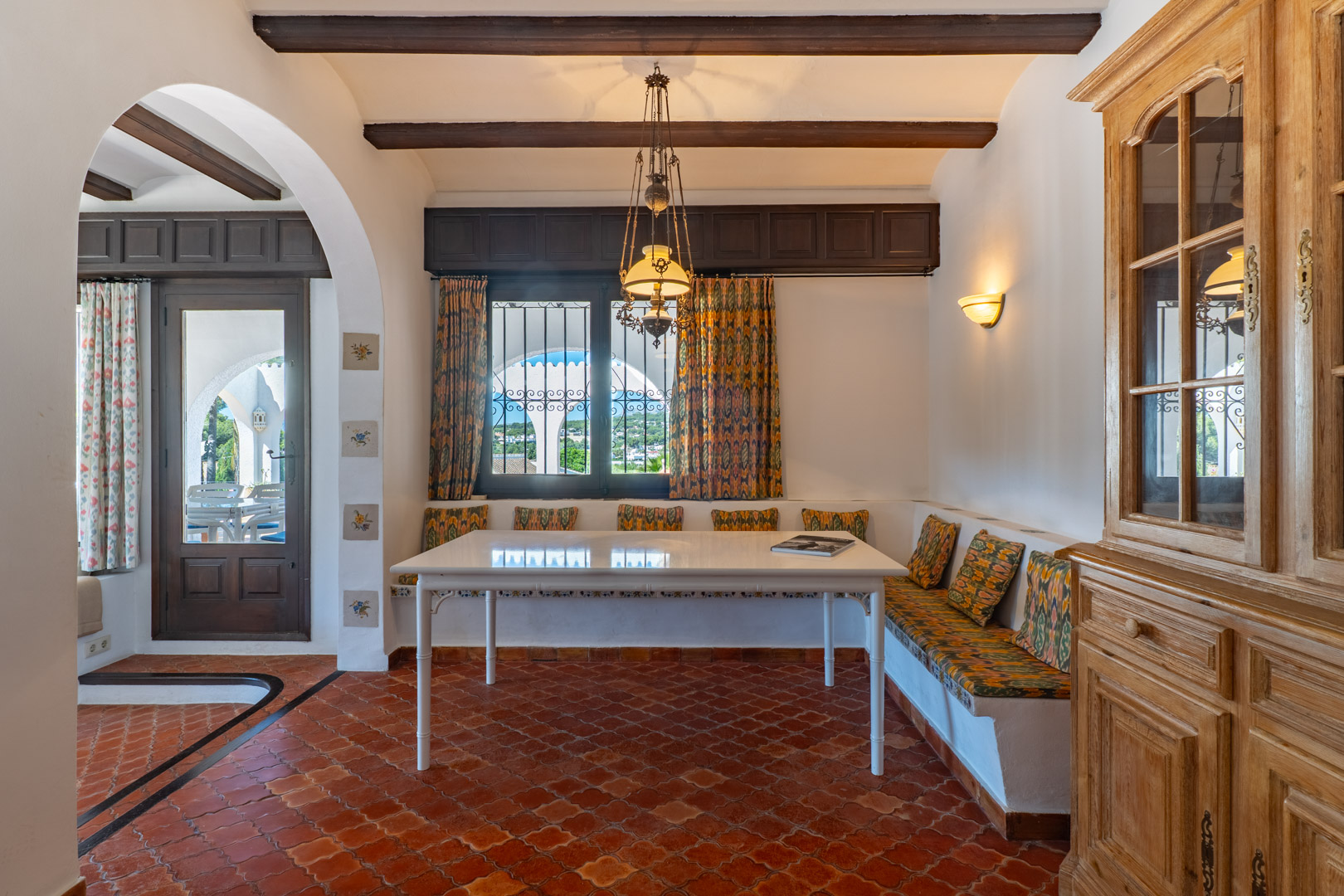 Eetkamer met ingebouwde bank, witte tafel en terracotta tegelvloer, verlicht door een kroonluchter.