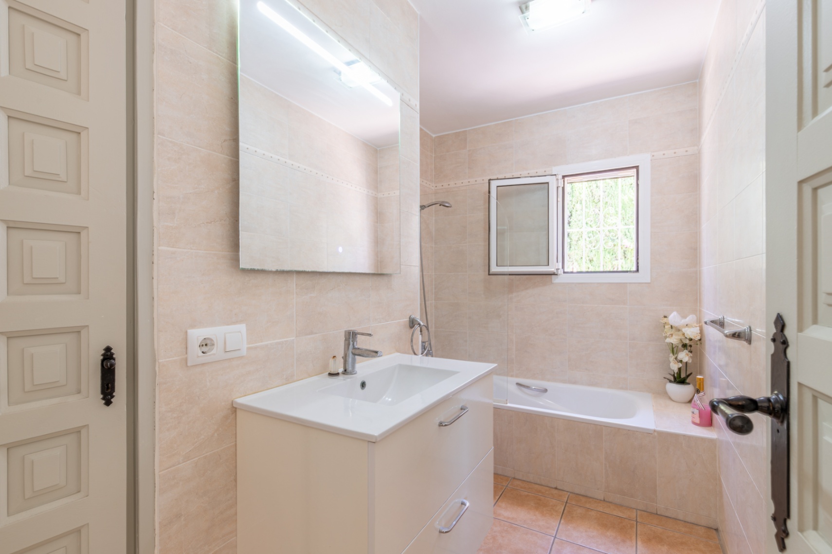 Badkamer met lichtbeige tegels, een wit wastafelmeubel en grote spiegel met bovenverlichting. Voorzien van een bad en raam voor natuurlijk lichtinval.