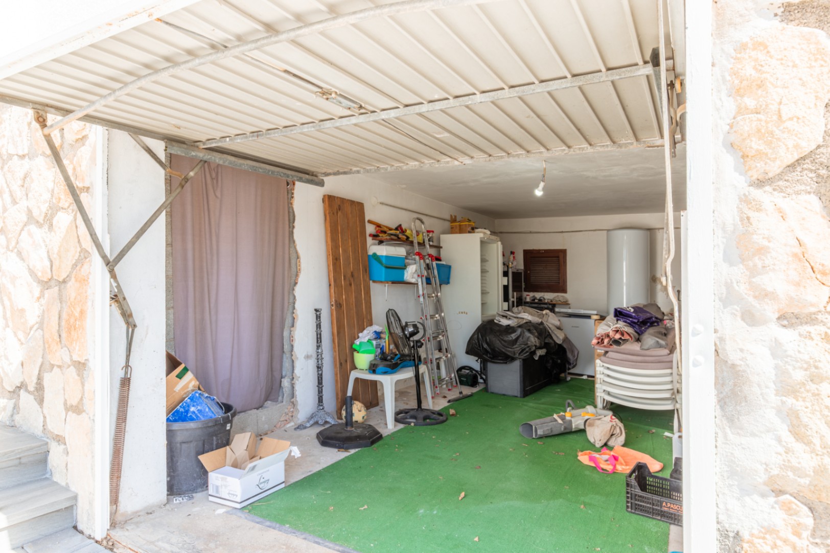 Garage- of opslagruimte met een golfplaten dak en groene kunstgrasvloer. Bevat een boiler, koelkast, ladder en diverse opgeslagen voorwerpen.