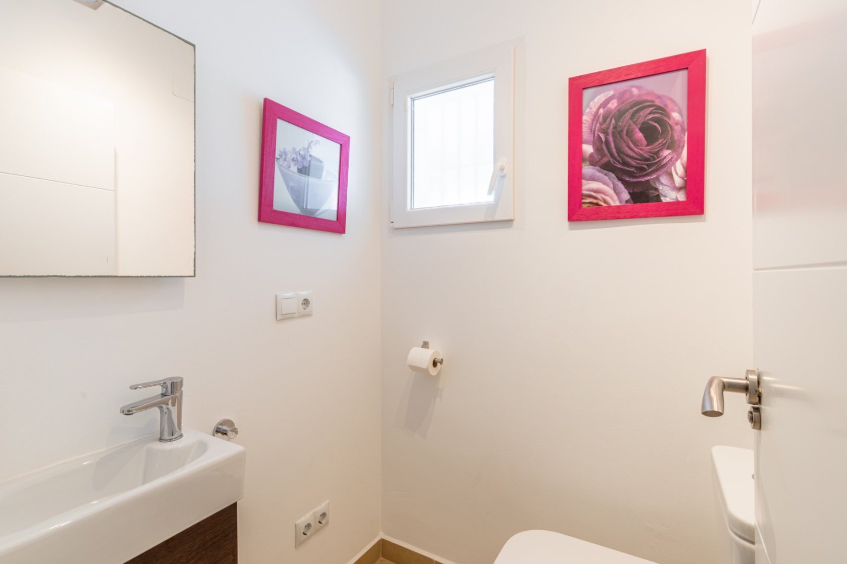 Gastentoilet met rechthoekige wastafel, moderne chromen kraan en donker houtlook meubel. Een klein raam zorgt voor natuurlijk licht, aangevuld met roze ingelijste kunst.