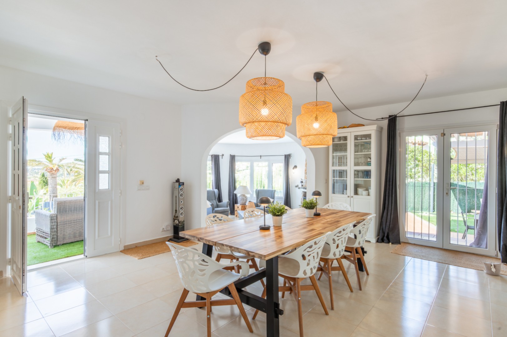 Lichte eetkamer met robuuste houten tafel, opvallende rotan hanglampen en tegelvloer. Directe toegang tot een privéterras en verbinding via een boog met de woonkamer.