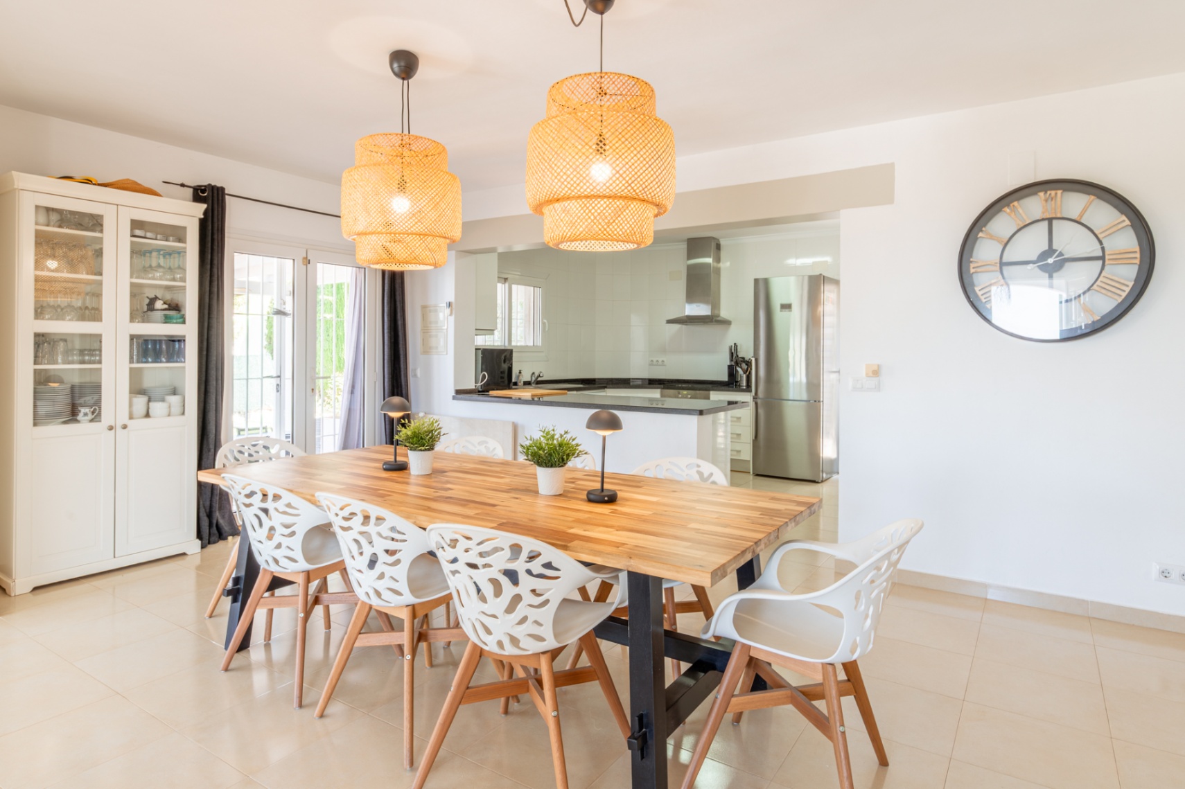 Lichte eetruimte met een grote houten tafel, acht kenmerkende witte stoelen en twee gevlochten hanglampen. Deze sluit naadloos aan op een moderne keuken met roestvrijstalen apparatuur.
