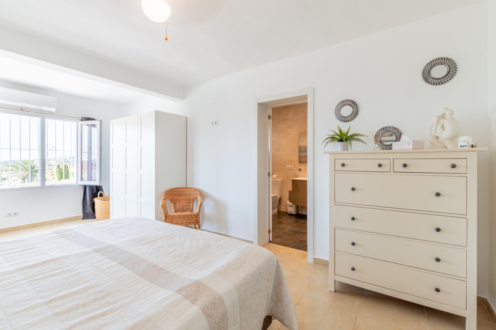 Lichte hoofdslaapkamer met tweepersoonsbed, witte kledingkast en directe toegang tot een en-suite badkamer. Het raam biedt uitzicht op de vegetatie.