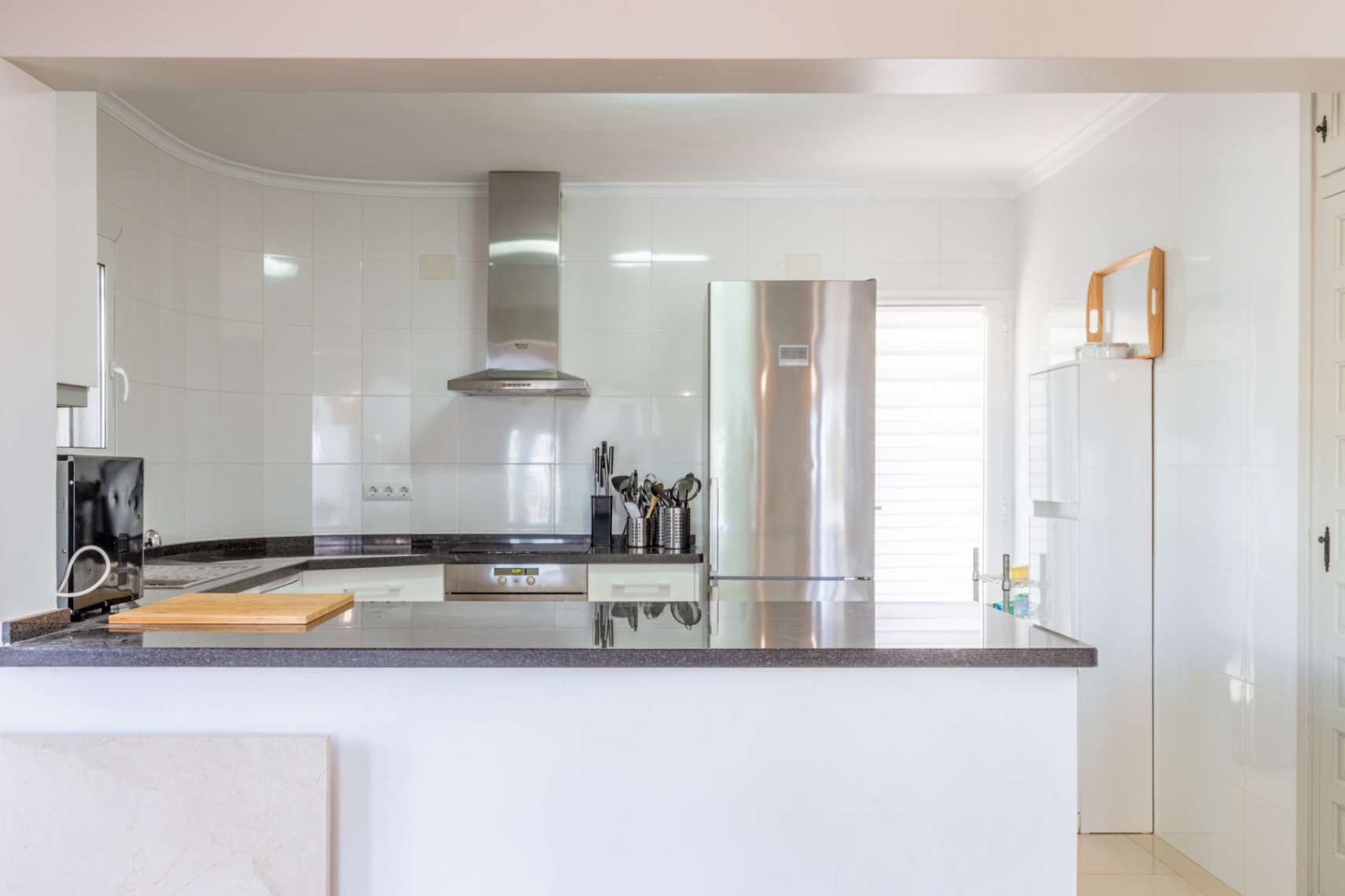 Lichte, moderne keuken met witte tegelwanden, donkere granieten werkbladen en geïntegreerde roestvrijstalen apparatuur, waaronder koelkast en afzuigkap. Een open balie biedt werkruimte.