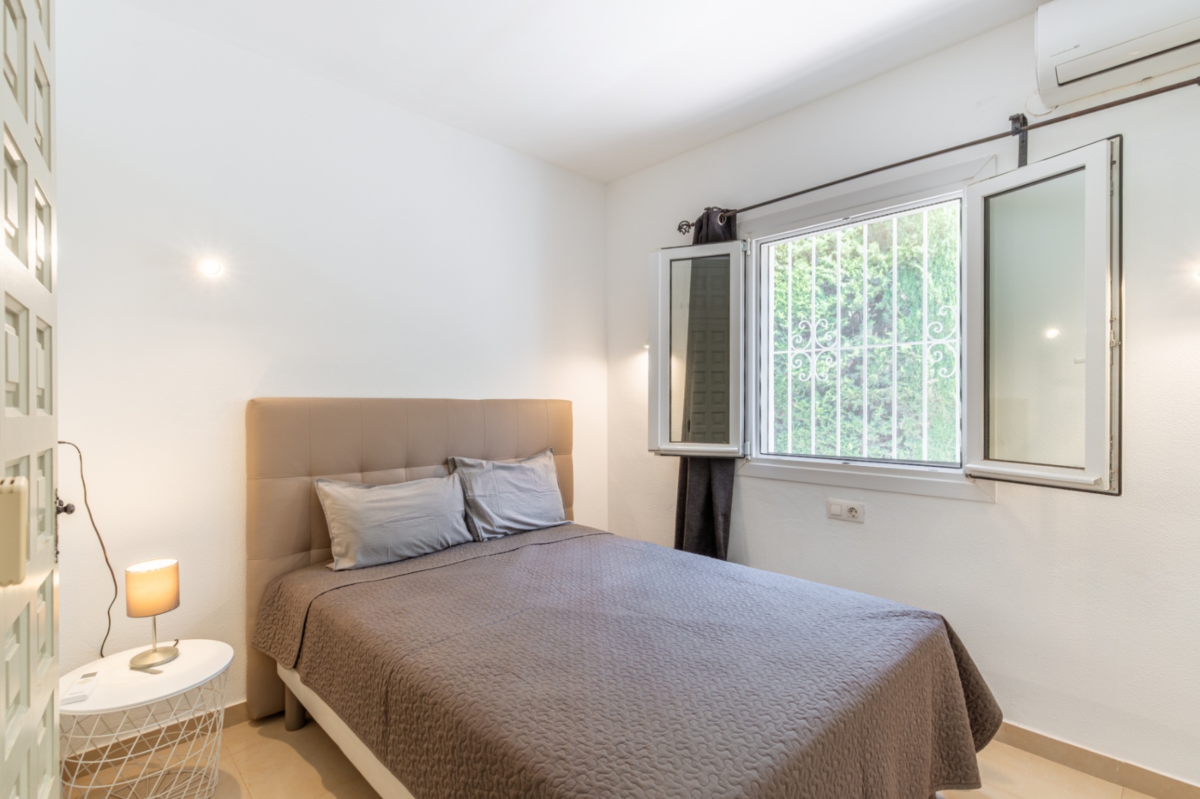 Lichte slaapkamer met comfortabel bed en gestoffeerd hoofdeinde, groot raam met veiligheidsroosters en uitzicht op groen, plus airconditioning.
