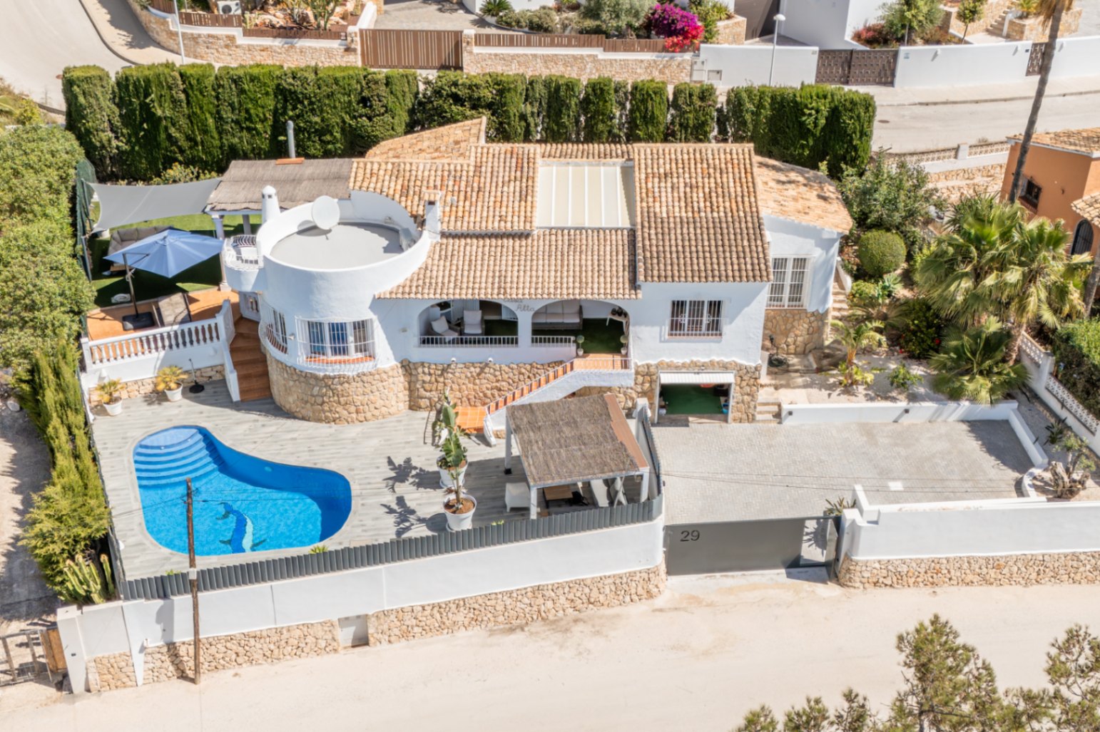 Luchtfoto van de witte villa met terracotta dak en niervormig privézwembad met dolfijnmozaïek. De woning beschikt over een ruime geplaveide oprit en diverse buitenterrassen.