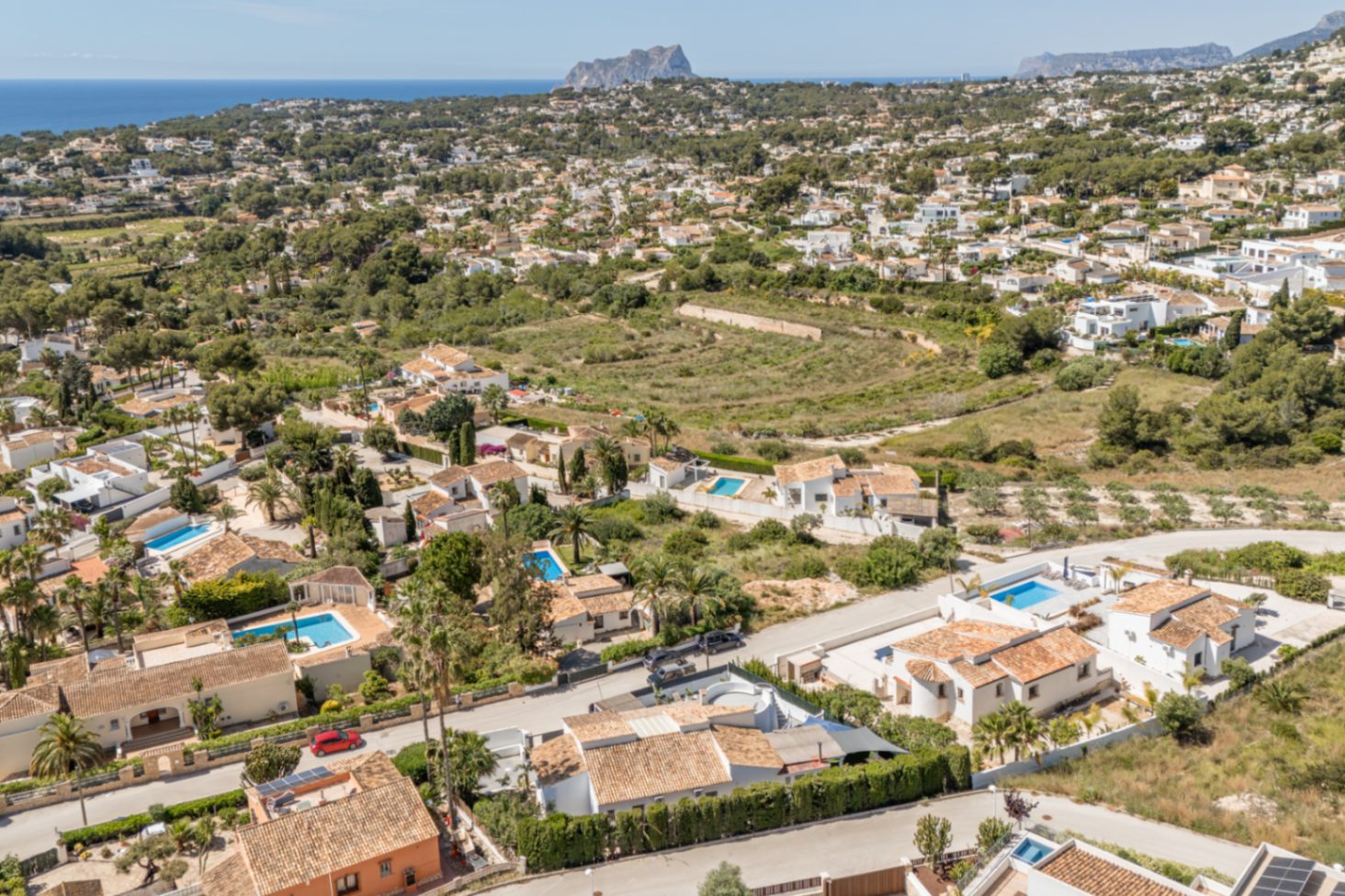 Luchtfoto van een woonwijk in Moraira, met diverse villa's met terracotta daken, privézwembaden en weelderig groen. De Middellandse Zee en een verre rotsachtige kaap omlijsten het kustlandschap.