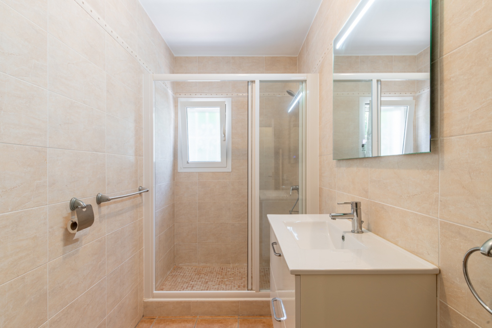 Moderne badkamer met beige wandtegels, een wit badmeubel met chromen kraan, een LED-verlichte spiegel en een inloopdouche met mozaïekvloer en raam.