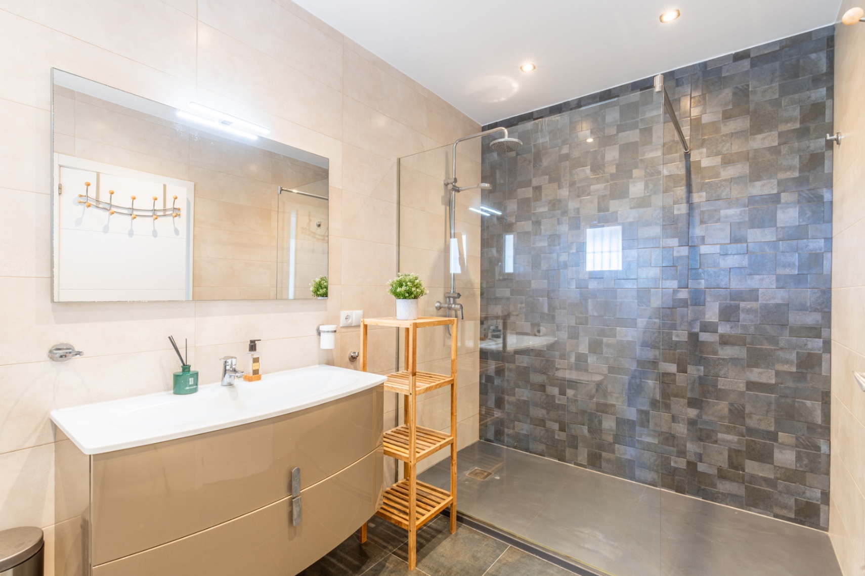 Moderne badkamer met glanzend beige wastafelmeubel, verlichte spiegel en inloopdouche met glazen wand en donkergrijze mozaïektegels. Lichte wandtegels, donkere vloer.