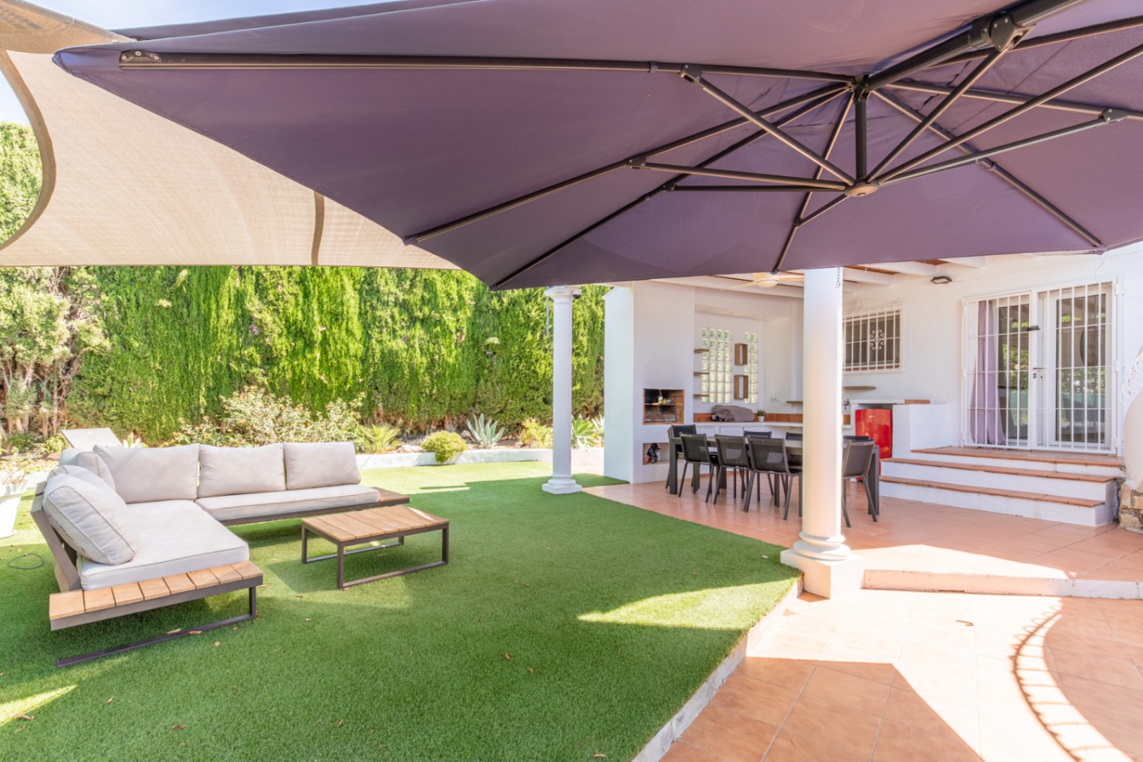Overdekt buitenterras met eethoek en ingebouwde BBQ, grenzend aan een kunstgrasveld met loungeset en een grote parasol voor schaduw.