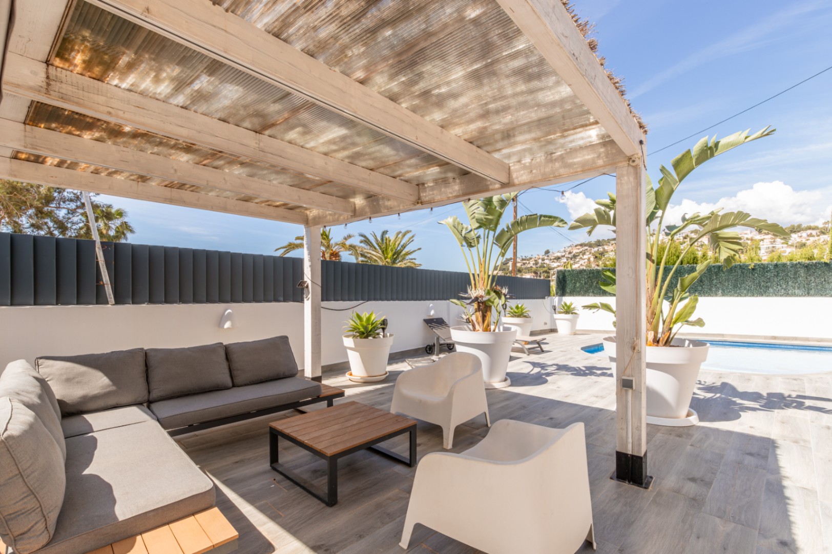 Overdekt buitenterras met houten pergola en doorschijnend dak, ingericht met een grijze modulaire bank, salontafel en tropische planten. Grijze tegelvloer met een glimp van het zwembad.