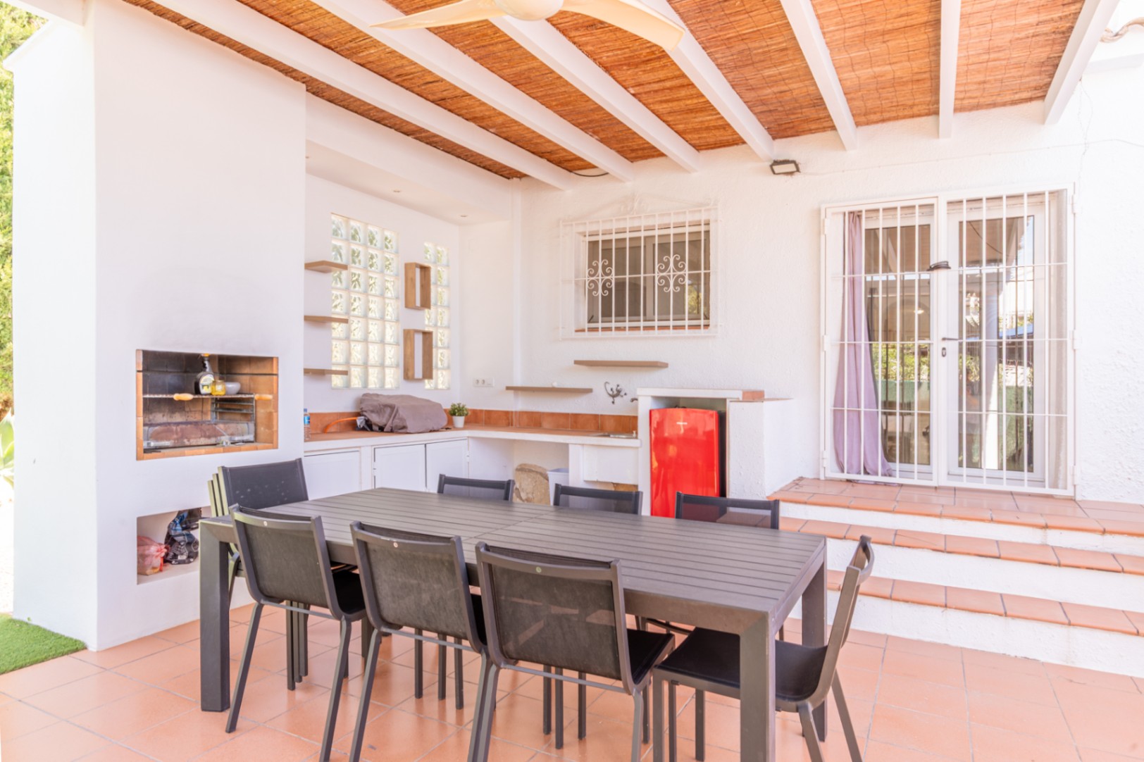 Overdekt buitenterras met ingebouwde barbecue, functionele buitenkeuken met rode minikoelkast en een grote eettafel. De ruimte is voorzien van terracottategels en een lattenplafond.