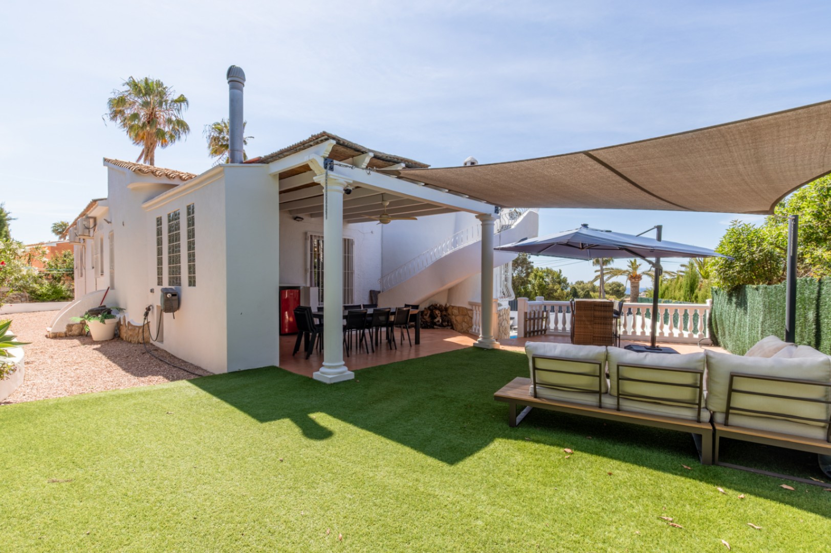 Ruim kunstgras gazon met een moderne buitenbank. Overdekt eetterras met wenteltrap, beschut onder een elegant schaduwdoek.