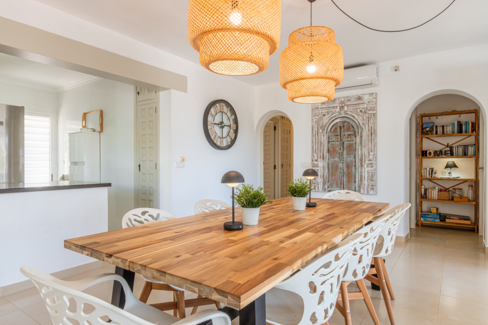 Ruime eetkamer met een robuuste houten tafel, moderne witte stoelen en twee geweven hanglampen. Bogen verbinden met de keuken en een leeshoek met houten boekenkast.