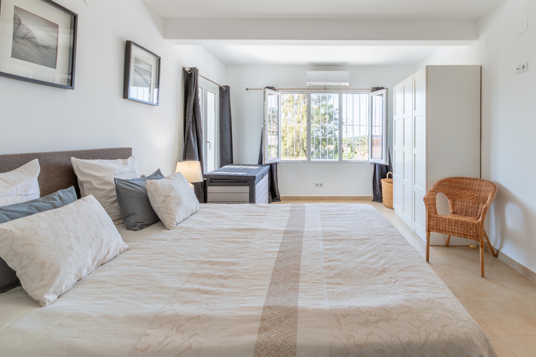 Ruime slaapkamer met groot bed, lichte tegelvloer, witte kledingkast en raam met tralies dat uitkijkt op groen, voorzien van airconditioning.