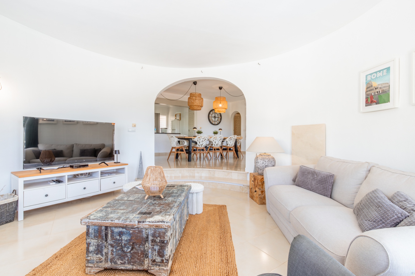 Ruime woonkamer met lichte tegels, comfortabele bank en rustieke houten salontafel op een jute kleed. Een boog leidt naar de verhoogde eetkamer met moderne stoelen en rotan hanglampen.