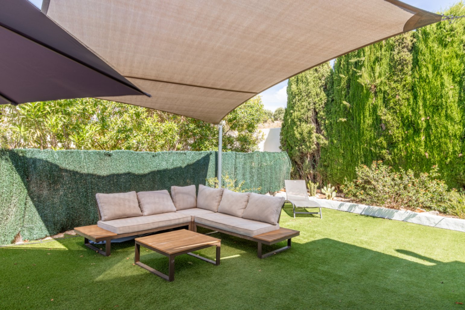 Schaduwrijke buitenlounge met L-vormige bank, lichte kussens en houten salontafel op kunstgras. Hoge heggen en bomen bieden privacy onder een groot beige schaduwdoek.