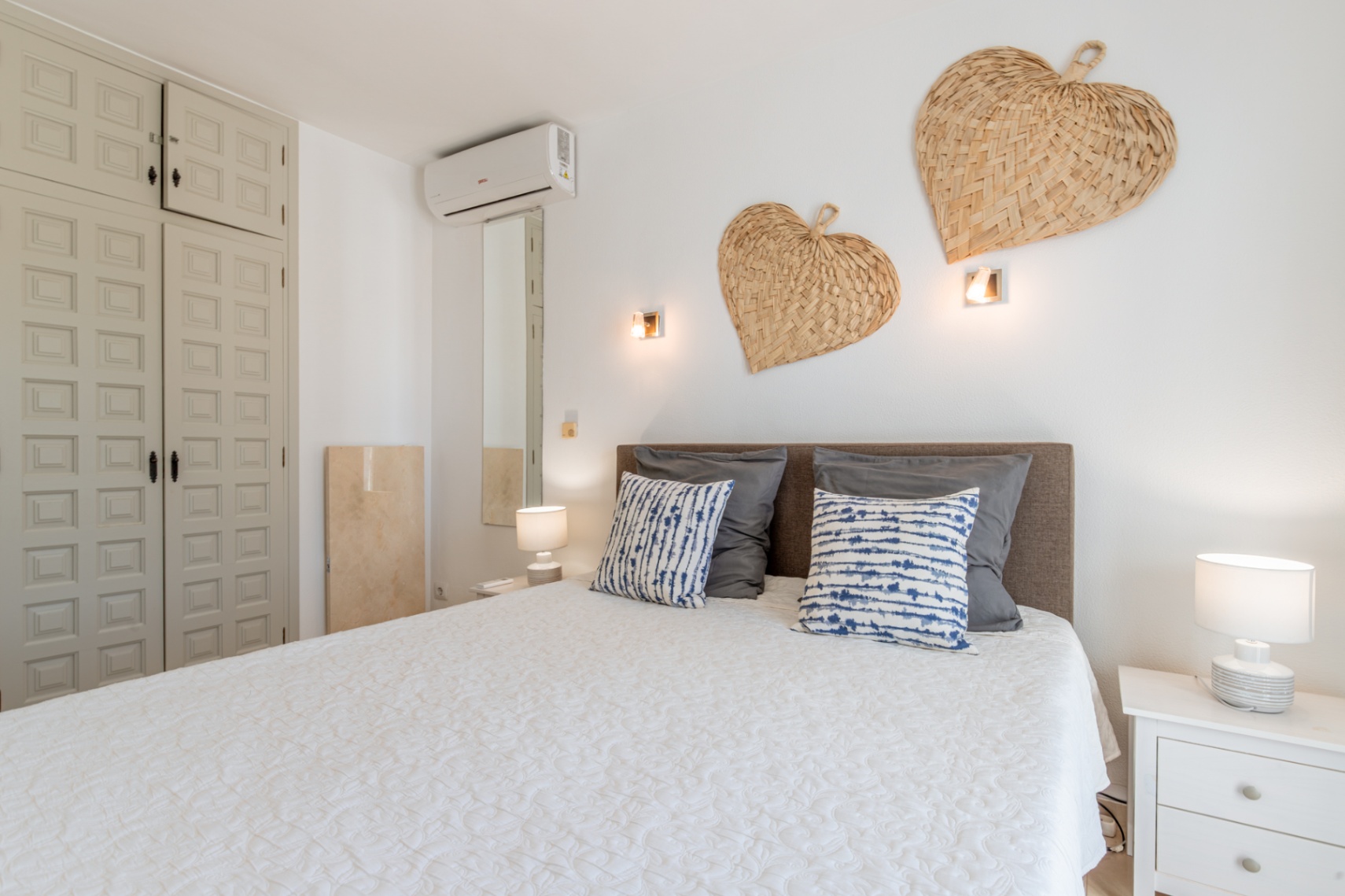 Slaapkamer met comfortabel bed, grijs hoofdbord, wit gewatteerd dekbed en kussens met patroon. Ingebouwde paneelkast, wandairconditioning en decoratieve gevlochten harten.