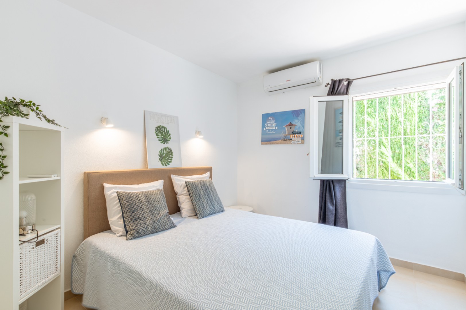 Slaapkamer met tweepersoonsbed, beige stoffen hoofdbord, airconditioning en een raam met veiligheidsrooster, uitkijkend op groen.