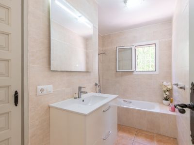 Badkamer met lichtbeige tegels, een wit wastafelmeubel en grote spiegel met bovenverlichting. Voorzien van een bad en raam voor natuurlijk lichtinval.