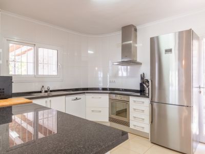 Keuken met witte kasten, donkere granieten werkbladen en roestvrijstalen apparatuur, waaronder een koelkast en inbouw oven. Raam met tralies zorgt voor natuurlijk licht.