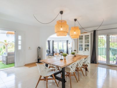 Lichte eetkamer met robuuste houten tafel, opvallende rotan hanglampen en tegelvloer. Directe toegang tot een privéterras en verbinding via een boog met de woonkamer.
