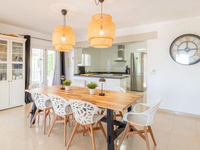 Lichte eetruimte met een grote houten tafel, acht kenmerkende witte stoelen en twee gevlochten hanglampen. Deze sluit naadloos aan op een moderne keuken met roestvrijstalen apparatuur.