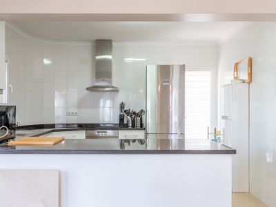 Lichte, moderne keuken met witte tegelwanden, donkere granieten werkbladen en geïntegreerde roestvrijstalen apparatuur, waaronder koelkast en afzuigkap. Een open balie biedt werkruimte.