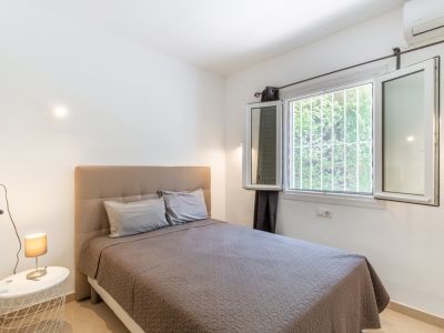 Lichte slaapkamer met comfortabel bed en gestoffeerd hoofdeinde, groot raam met veiligheidsroosters en uitzicht op groen, plus airconditioning.