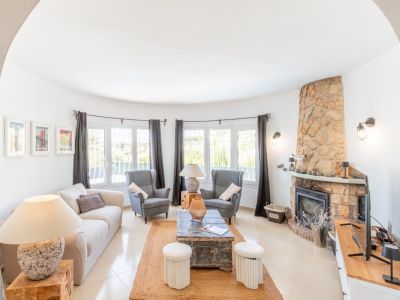 Lichte woonkamer met een gebogen raamwand voor natuurlijk licht, een opvallende stenen open haard en lichte tegelvloeren. Een jute vloerkleed omkadert de zithoek.