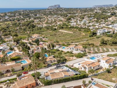 Luchtfoto van een woonwijk in Moraira, met diverse villa's met terracotta daken, privézwembaden en weelderig groen. De Middellandse Zee en een verre rotsachtige kaap omlijsten het kustlandschap.