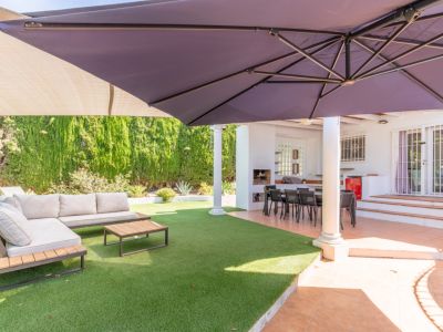 Overdekt buitenterras met eethoek en ingebouwde BBQ, grenzend aan een kunstgrasveld met loungeset en een grote parasol voor schaduw.