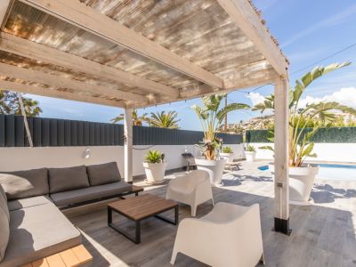 Overdekt buitenterras met houten pergola en doorschijnend dak, ingericht met een grijze modulaire bank, salontafel en tropische planten. Grijze tegelvloer met een glimp van het zwembad.