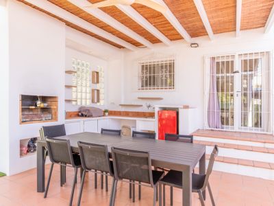Overdekt buitenterras met ingebouwde barbecue, functionele buitenkeuken met rode minikoelkast en een grote eettafel. De ruimte is voorzien van terracottategels en een lattenplafond.