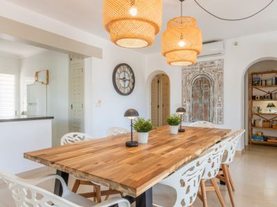 Ruime eetkamer met een robuuste houten tafel, moderne witte stoelen en twee geweven hanglampen. Bogen verbinden met de keuken en een leeshoek met houten boekenkast.