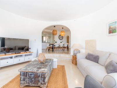 Ruime woonkamer met lichte tegels, comfortabele bank en rustieke houten salontafel op een jute kleed. Een boog leidt naar de verhoogde eetkamer met moderne stoelen en rotan hanglampen.