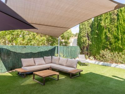 Schaduwrijke buitenlounge met L-vormige bank, lichte kussens en houten salontafel op kunstgras. Hoge heggen en bomen bieden privacy onder een groot beige schaduwdoek.