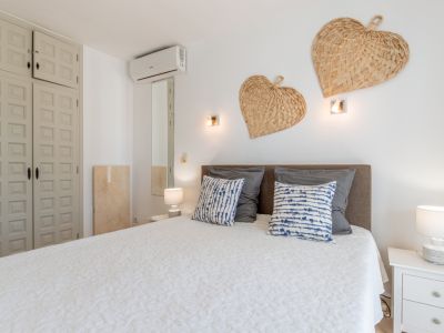 Slaapkamer met comfortabel bed, grijs hoofdbord, wit gewatteerd dekbed en kussens met patroon. Ingebouwde paneelkast, wandairconditioning en decoratieve gevlochten harten.