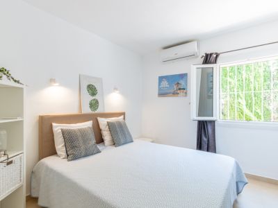 Slaapkamer met tweepersoonsbed, beige stoffen hoofdbord, airconditioning en een raam met veiligheidsrooster, uitkijkend op groen.
