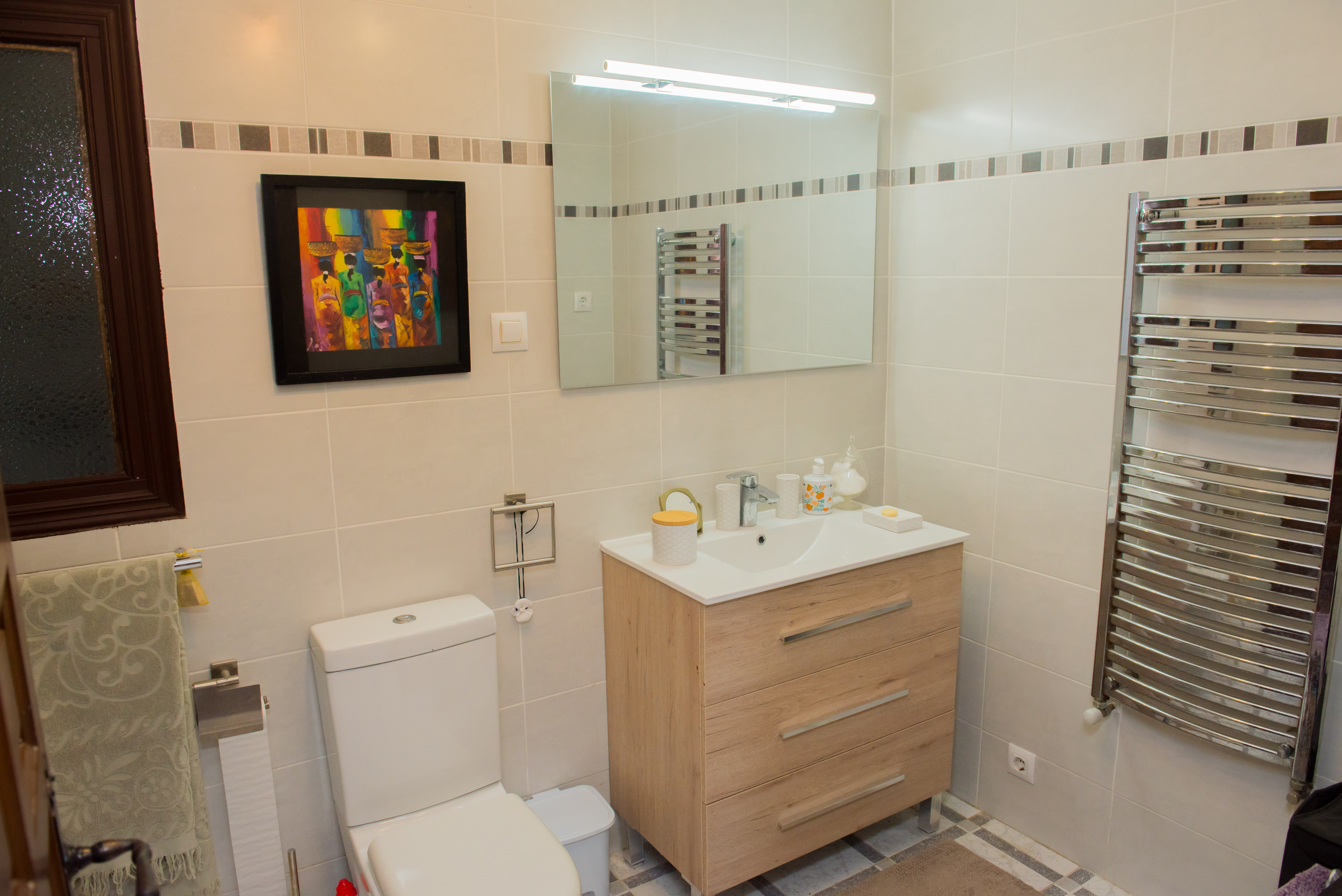 Badkamer met crèmekleurige tegels, licht houten wastafelmeubel en een verchroomd handdoekenrek.