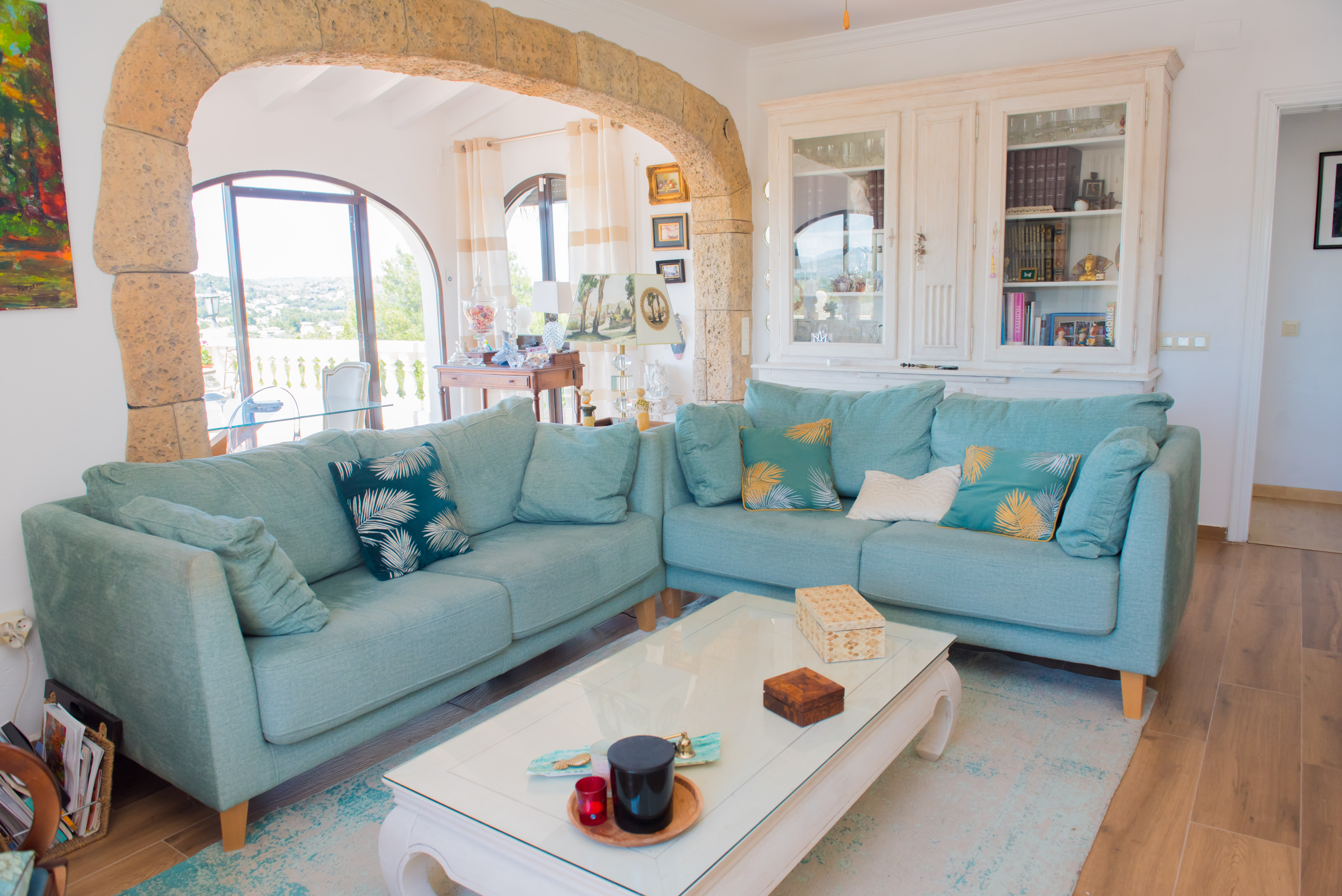 Lichte woonkamer met turquoise hoekbank, witte salontafel en rustieke stenen boog die uitzicht biedt op de buitenomgeving.