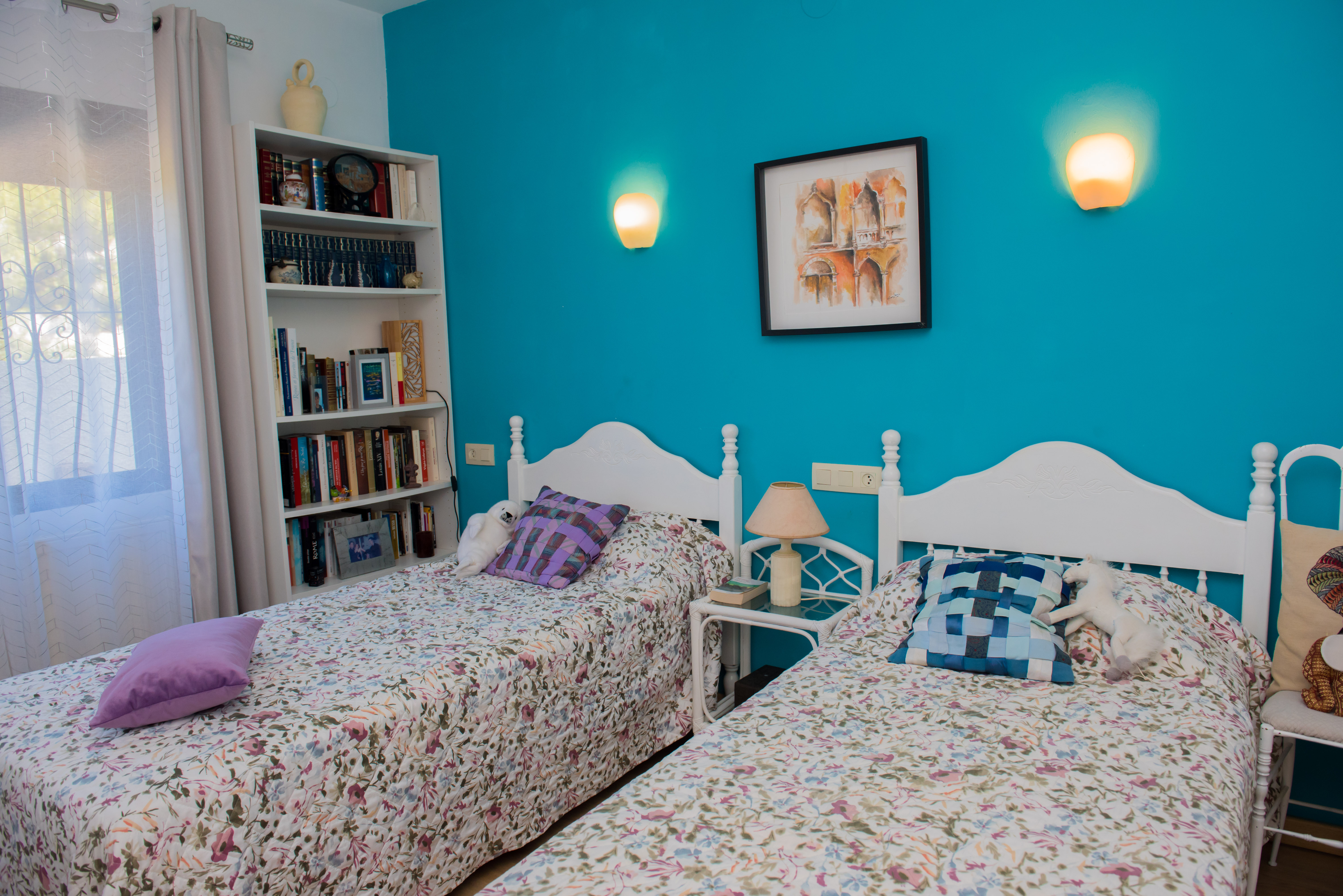 Slaapkamer met twee eenpersoonsbedden, turquoise muur en een witte boekenkast gevuld met boeken.