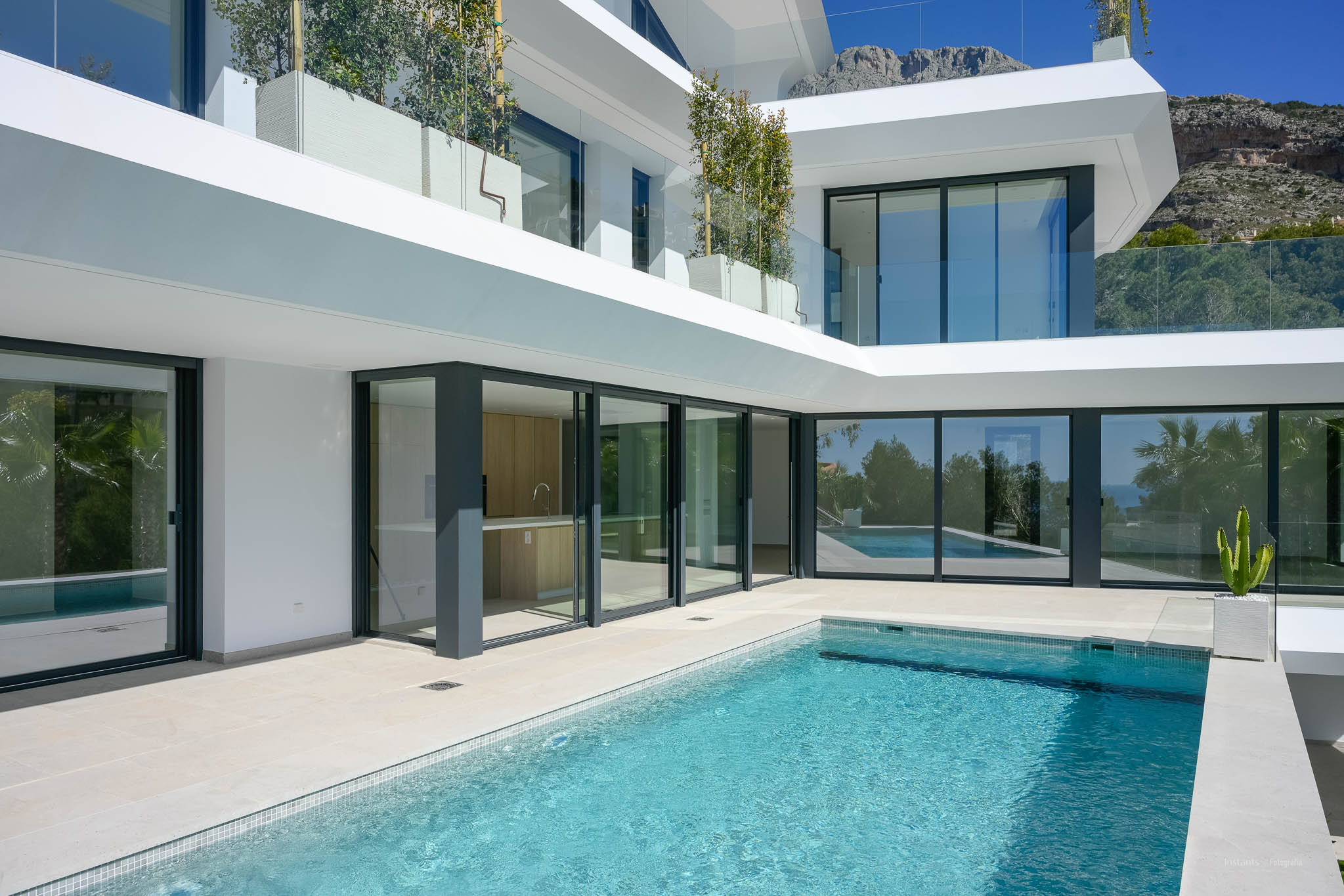 Buitenbad met lichte tegels en beige terras, geflankeerd door grote glazen puien die een buitenkeuken en het villa-interieur tonen, met uitzicht op de bergen.