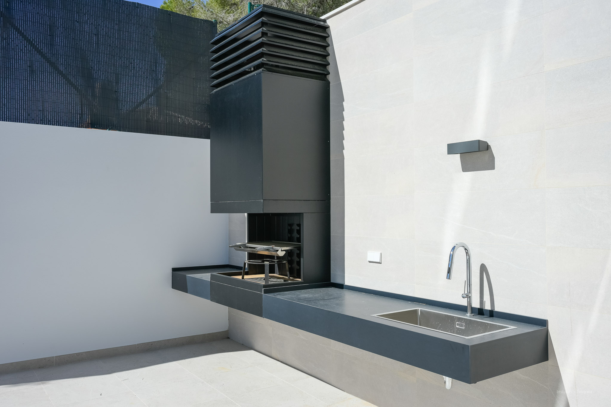 Buitenkeuken met geïntegreerde donkergrijze barbecue, een bijpassend aanrechtblad en een roestvrijstalen spoelbak. Een moderne chromen kraan completeert de lichte tegelwanden.