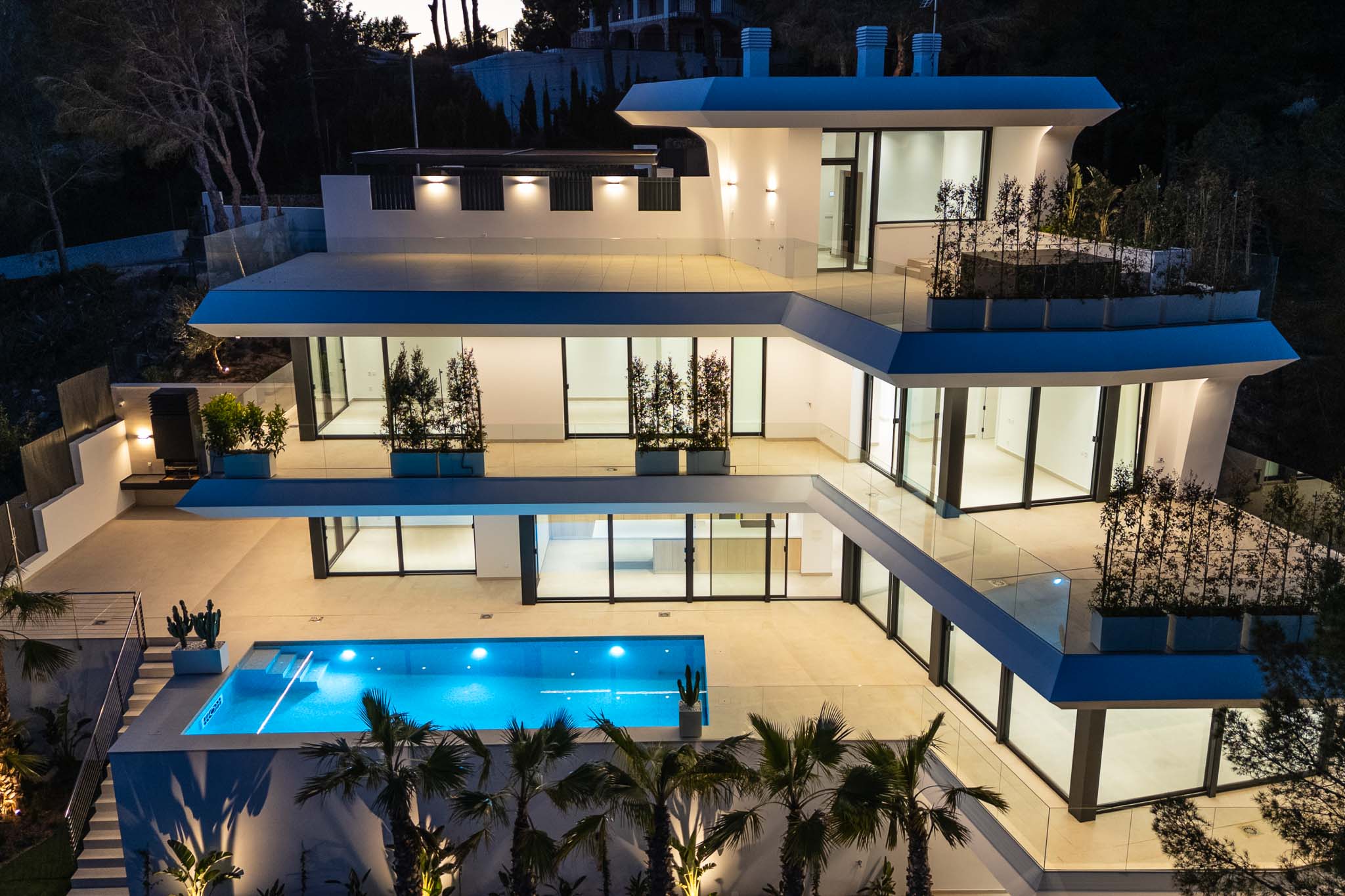 De meerlaagse moderne architectuur van de villa licht op in de schemering, met een levendig blauw zwembad en ruime terrassen met glazen balustrades.